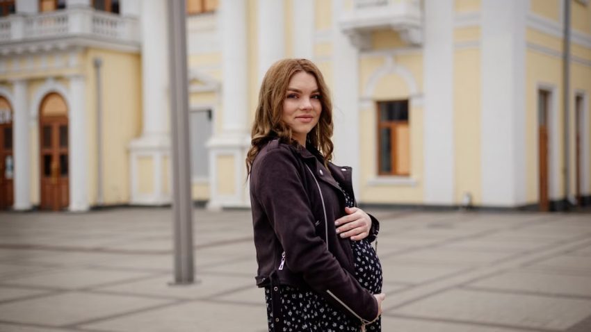 A pregnant woman in a black jacket stands smiling in an urban environment.